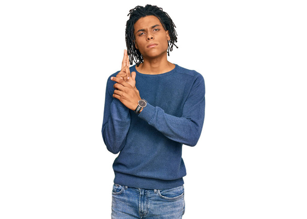 Joven afroamericano vestido con suéter casual de invierno sosteniendo arma simbólica con gesto de mano, jugando a matar armas de fuego, cara enojada  - Foto, imagen