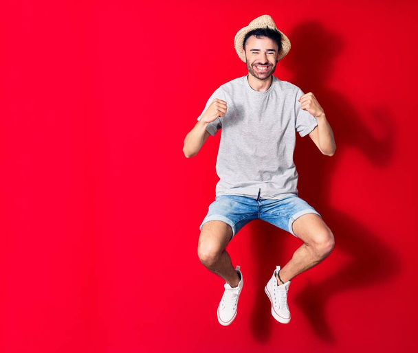 Young handsome hispanic man wearing casual clothes and summer hat smiling happy. Jumping with smile on face celebrating with fists up over isolated red background. - Φωτογραφία, εικόνα