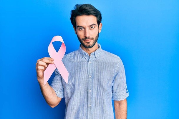 Joven hombre hispano sosteniendo actitud de pensamiento de cinta de cáncer rosa y expresión sobria buscando confianza en sí mismo  - Foto, Imagen