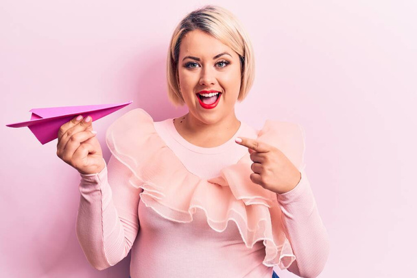 Young beautiful blonde plus size woman holding paper airplane over isolated pink background smiling happy pointing with hand and finger - Fotografie, Obrázek