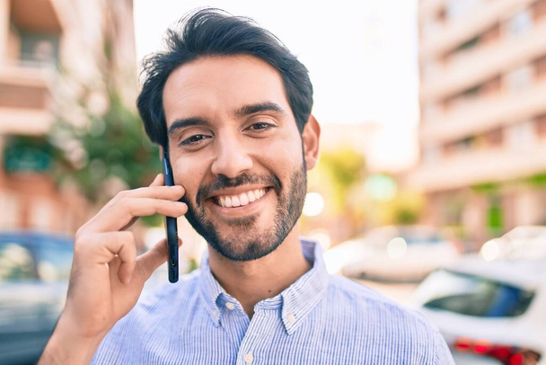Joven hombre hispano sonriendo feliz hablando en el teléfono inteligente en la ciudad. - Foto, Imagen