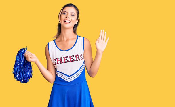 Giovane bella donna indossa uniforme cheerleader rinuncia dicendo ciao felice e sorridente, amichevole gesto di benvenuto  - Foto, immagini