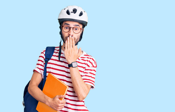 Młody Hiszpan noszący plecak studencki i kask rowerowy trzymający segregator zakrywający usta dłonią, zszokowany i bojący się pomyłki. zaskoczona ekspresja  - Zdjęcie, obraz