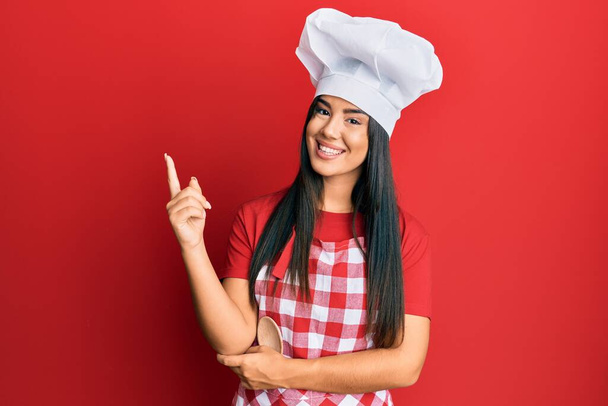 Mladá krásná hispánská dívka v pekařské uniformě a kuchař klobouk s úsměvem šťastný ukazující rukou a prstem na stranu  - Fotografie, Obrázek
