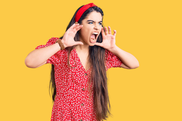Μπρουνέτ έφηβο κορίτσι φορώντας καλοκαιρινό φόρεμα φωνάζοντας θυμωμένος δυνατά με τα χέρια πάνω από το στόμα  - Φωτογραφία, εικόνα