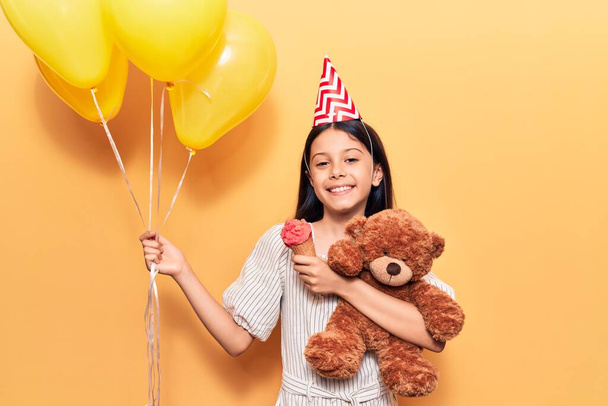 Αξιολάτρευτο ισπανόφωνο κορίτσι που φοράει καπέλο γενεθλίων χαμογελώντας χαρούμενο. Μόνιμη με χαμόγελο στο πρόσωπο κρατώντας μπαλόνια και αρκουδάκι τρώει παγωτό πάνω από απομονωμένο κίτρινο φόντο - Φωτογραφία, εικόνα