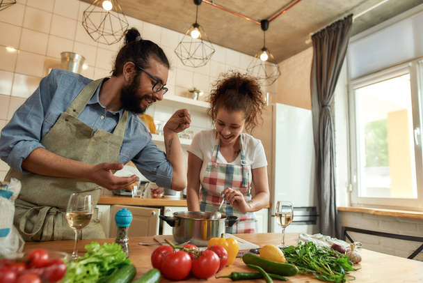 Italian man adding pepper, spice to the soup while woman stirring it and smiling. Couple preparing a meal together in the kitchen. Cooking at home, Italian cuisine - Foto, Imagem