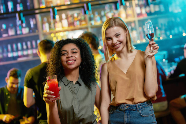 Attractive young women smiling at camera, posing with cocktail in their hands. Friends celebrating, having fun in the bar - Zdjęcie, obraz
