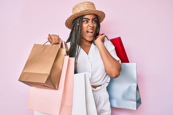 Young african american woman with braids holding shopping bags angry and mad screaming frustrated and furious, shouting with anger. rage and aggressive concept.  - Foto, imagen