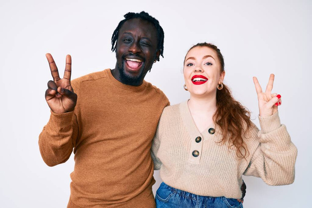 Interracial couple wearing casual clothes smiling with happy face winking at the camera doing victory sign with fingers. number two.  - Photo, Image