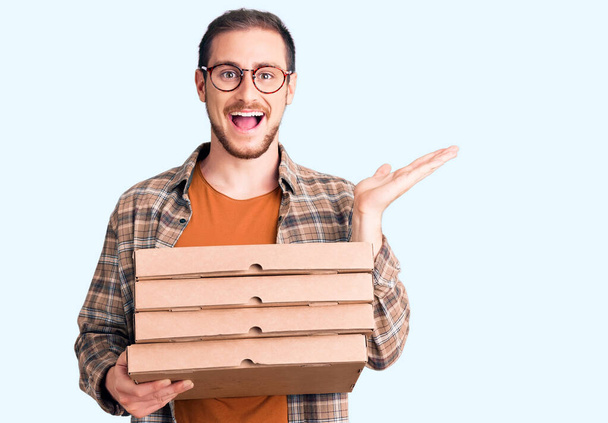 Joven hombre caucásico guapo sosteniendo la caja de pizza de entrega celebrando la victoria con sonrisa feliz y la expresión ganadora con las manos levantadas  - Foto, imagen