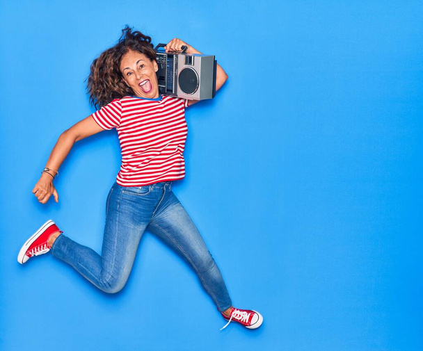 Middle age beautiful woman smiling happy. Jumping with smile on face listening to music using boombox over isolated blue background - Fotó, kép