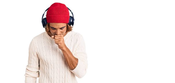 Jonge Spaanse man die naar muziek luistert met een koptelefoon die zich onwel voelt en hoest als symptoom voor verkoudheid of bronchitis. Gezondheidszorgconcept.  - Foto, afbeelding