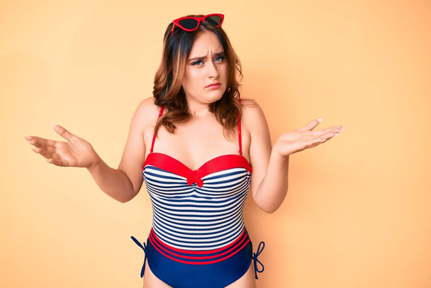 Young beautiful caucasian woman wearing swimsuit and sunglasses clueless and confused expression with arms and hands raised. doubt concept.  - Photo, Image