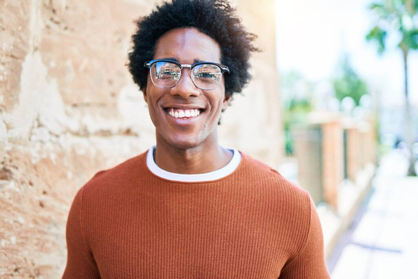Mladý pohledný africký Američan v neformálním oblečení a brýlích se šťastně usmívá. Stojící s úsměvem na tváři dívající se na kameru kráčející po ulici. - Fotografie, Obrázek