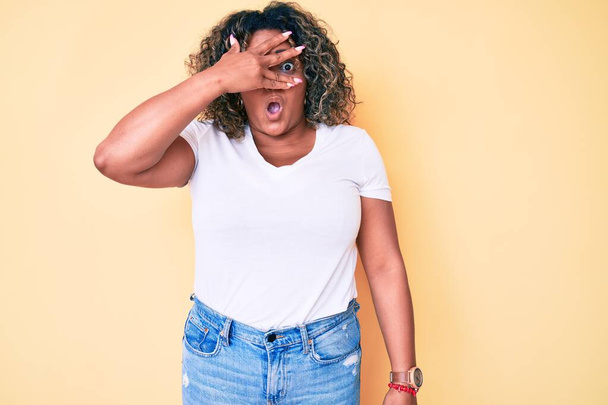 Joven afroamericana mujer de talla grande vistiendo camiseta blanca casual mirando con la mano a través de los dedos con expresión avergonzada.  - Foto, imagen