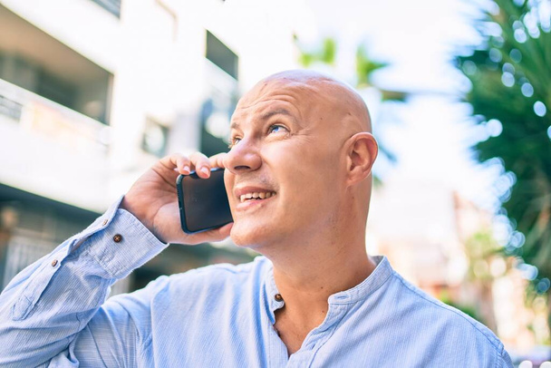 Homem careca de meia idade sorrindo feliz falando no smartphone na cidade. - Foto, Imagem
