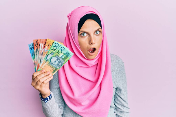 Joven mujer caucásica con hiyab islámico sosteniendo billetes de dólares australianos asustada y sorprendida con la boca abierta para sorpresa, cara de incredulidad  - Foto, imagen
