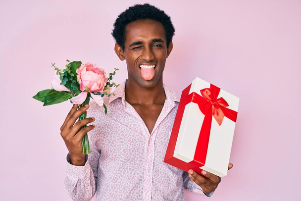 Αφρικανός όμορφος άντρας που κρατάει δώρο επετείου και μπουκέτο λουλούδια που βγάζουν τη γλώσσα του χαρούμενη με αστεία έκφραση.  - Φωτογραφία, εικόνα