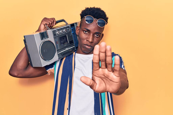 Młody Afroamerykanin trzyma boombox, słuchając muzyki z otwartą ręką robi znak stop z poważnym i pewnym siebie wyrazem twarzy, gest obrony  - Zdjęcie, obraz