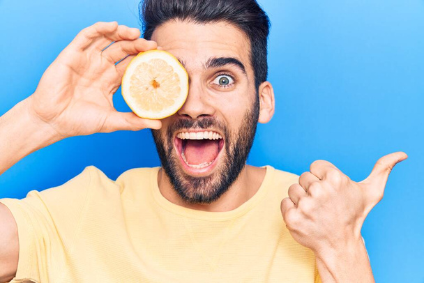 Молодий красивий чоловік з бородою тримає шматочок лимона над оком, вказуючи великий палець в сторону посміхаючись щасливим з відкритим ротом
  - Фото, зображення