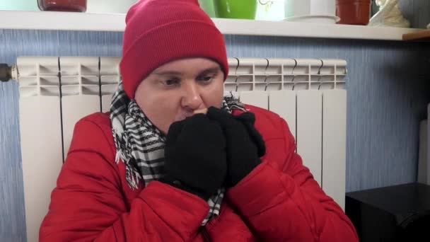 Άνδρας φορούν ζεστά ρούχα και πλεκτό καπάκι παγώσει στο σπίτι κάθεται κοντά στο κεντρικό σύστημα θέρμανσης - Πλάνα, βίντεο