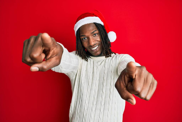 Afro-Américain avec des tresses portant un chapeau de Noël pointant vers vous et la caméra avec les doigts, souriant positif et joyeux  - Photo, image