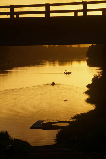 Rowers e barca di sicurezza sagomati sotto un ponte in un'alba d'oro sul fiume.                                - Foto, immagini