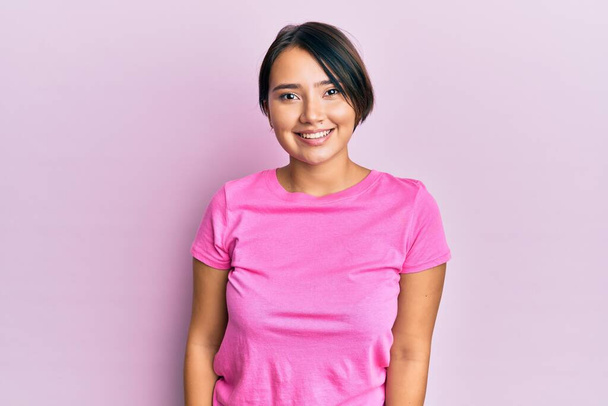 Mooie jonge vrouw met kort haar draagt roze tshirt over roze achtergrond met een vrolijke en koele glimlach op het gezicht. gelukkig persoon.  - Foto, afbeelding