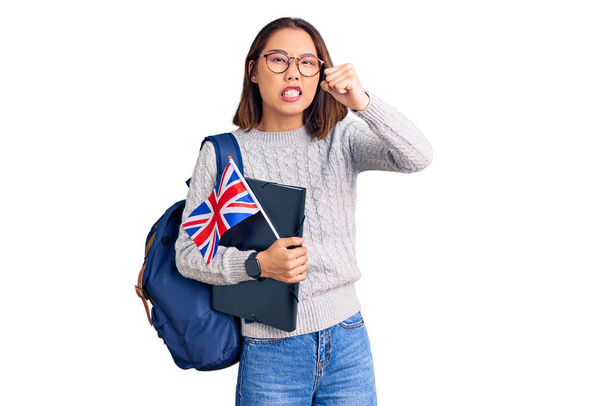 Fiatal gyönyörű kínai lány visel diák hátizsák gazdaság kötőanyag és Egyesült Királyság zászló bosszús és frusztrált kiabál a harag, kiabál őrült harag és kéz emelt  - Fotó, kép