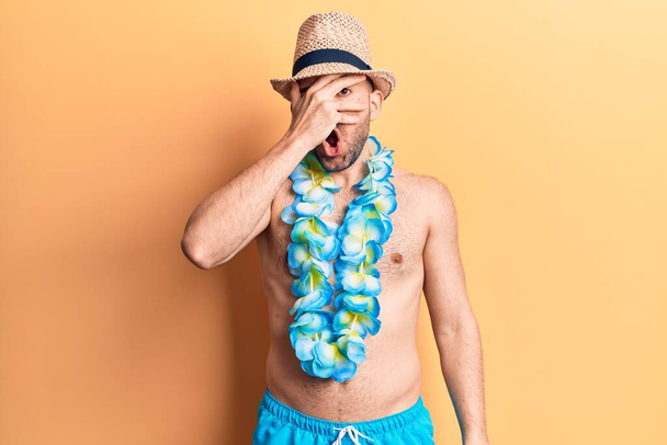Young handsome bald man shirtless wearing swimwear and hawaiian lei peeking in shock covering face and eyes with hand, looking through fingers afraid  - Fotoğraf, Görsel