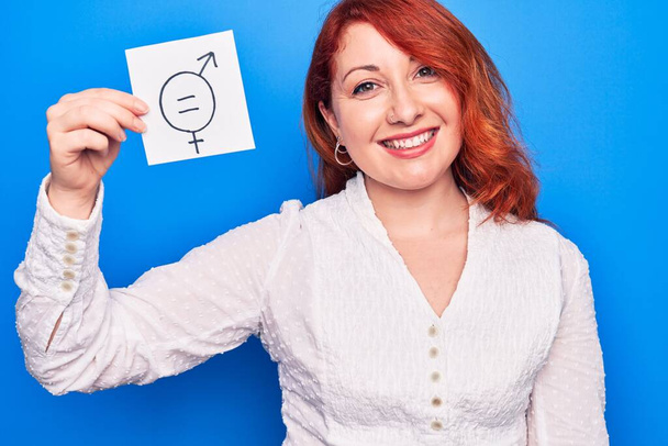 Молодая рыжая женщина с просьбой о дискриминации по половому признаку держит бумагу с сообщением о гендерном равенстве, выглядит позитивно и счастливо стоя и улыбаясь с уверенной улыбкой, показывая зубы - Фото, изображение