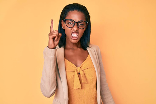 Giovane donna afroamericana con indosso abiti da lavoro che puntano il dito verso l'alto con l'idea di successo. Uscito e felice. numero uno.  - Foto, immagini