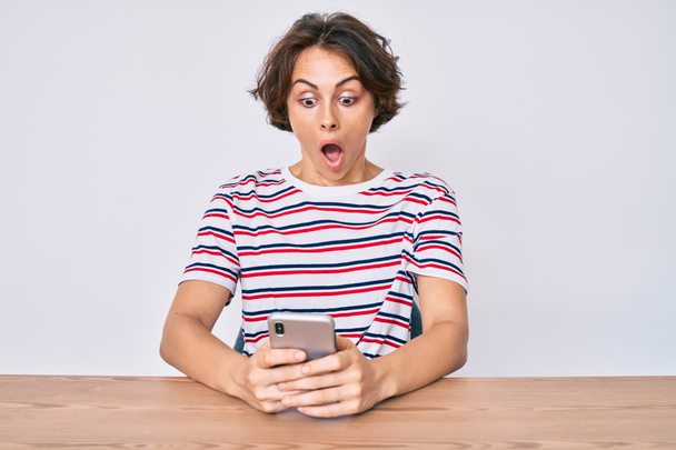 Young hispanic woman using smartphone sitting on the table scared and amazed with open mouth for surprise, disbelief face  - Photo, Image