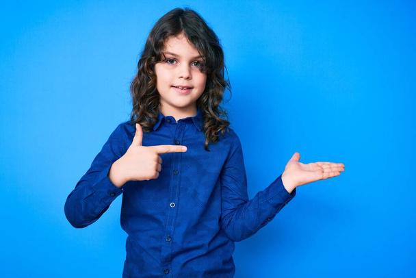 Schattig Latijns-Amerikaans kind met lang haar in casual kleding verbaasd en glimlachend naar de camera terwijl presenteren met de hand en wijzen met de vinger.  - Foto, afbeelding