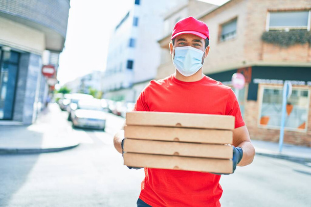 Młody dostawca w mundurze i masce medycznej ochrony przed koronawirusami. Trzymanie pudełek po pizzy na ulicy.. - Zdjęcie, obraz