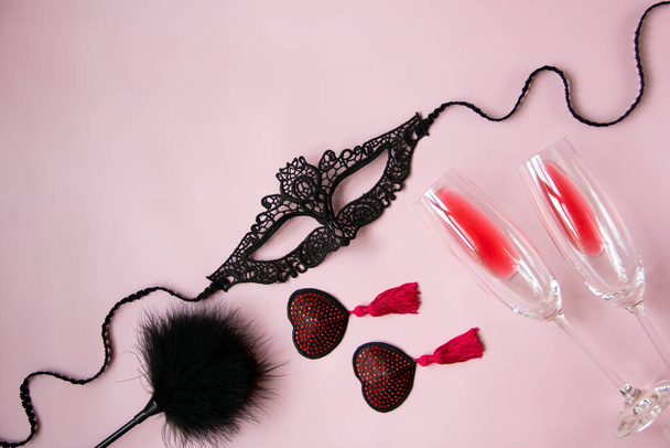 Una máscara de encaje para una mujer, borlas rojas empanadas burlescas, una paleta de plumas, copas de vino sobre un fondo rosa. Juguetes sexy para juegos para adultos. Piso tendido, vista superior. - Foto, imagen