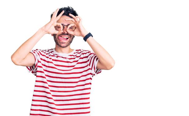 Un joven hispano vestido con ropa casual haciendo un buen gesto como binoculares sacando la lengua, ojos mirando a través de los dedos. expresión loca.  - Foto, Imagen