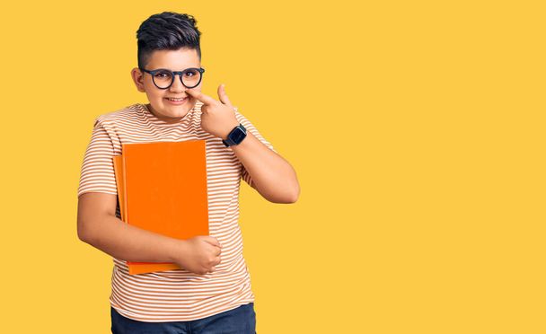 Menino menino segurando livro usando óculos apontando com a mão de dedo para cara e nariz, sorrindo alegre. conceito de beleza  - Foto, Imagem