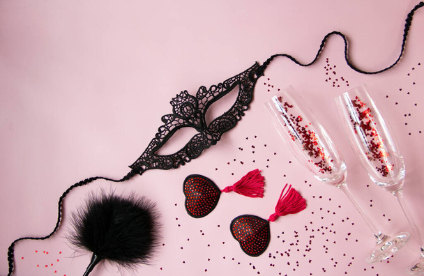 Μια δαντελένια μάσκα για μια γυναίκα, κόκκινες φούντες μπουρλέσκ, ένα φτερό κουπί, ποτήρια κρασί σε ροζ φόντο. Σέξι παιχνίδια για ενήλικες παιχνίδια. Επίπεδο lay, πάνω όψη. - Φωτογραφία, εικόνα