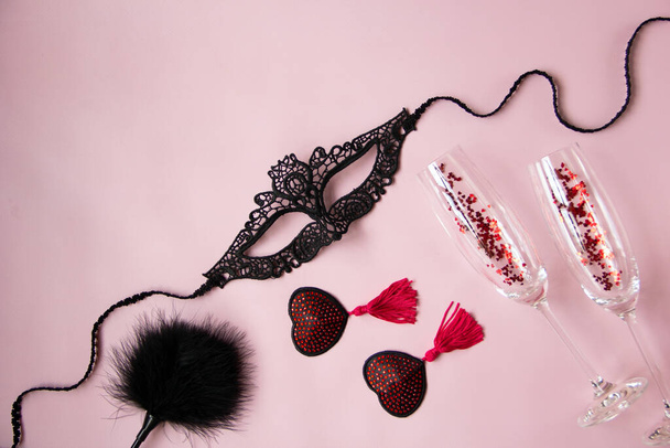Een kanten masker voor een vrouw, rode kwastjes burleske pasteitjes, een veer peddel, glazen wijn op een roze achtergrond. Sexy speelgoed voor volwassen spelletjes. Vlakke lay, bovenaanzicht. - Foto, afbeelding