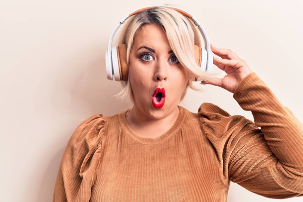 Beautiful blonde plus size woman listening to music using headphones over white background scared and amazed with open mouth for surprise, disbelief face - Photo, Image