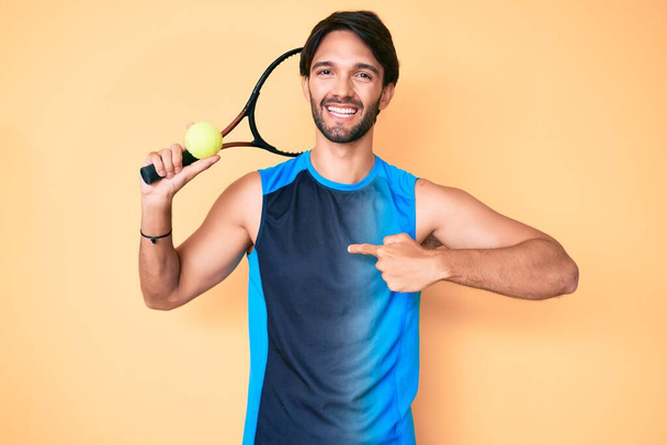 Hombre hispano guapo jugando tenis sosteniendo raqueta y pelota señalando con el dedo a uno mismo sonriendo feliz y orgulloso  - Foto, Imagen