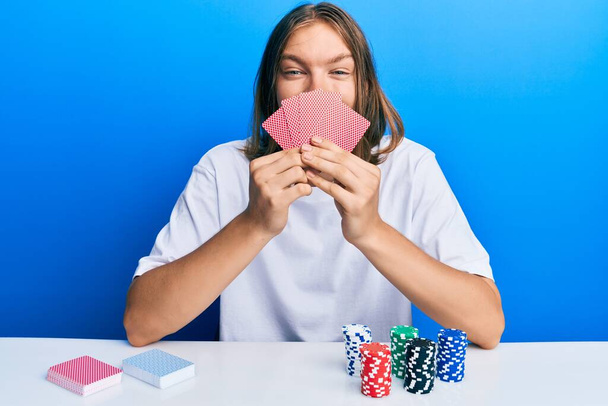 Przystojny biały mężczyzna z długimi włosami gra w pokera pokrywając twarz kartami uśmiechając się i śmiejąc głośno, bo śmieszne szalony żart.  - Zdjęcie, obraz