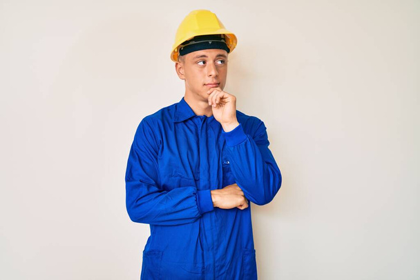 Young hispanic boy wearing worker uniform and hardhat serious face thinking about question with hand on chin, thoughtful about confusing idea  - Foto, Bild