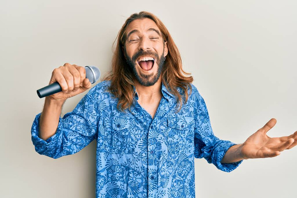 Atrakcyjny mężczyzna z długimi włosami i brodą śpiewający piosenkę za pomocą mikrofonu świętującego zwycięstwo szczęśliwym uśmiechem i zwycięską ekspresją z podniesionymi rękami  - Zdjęcie, obraz