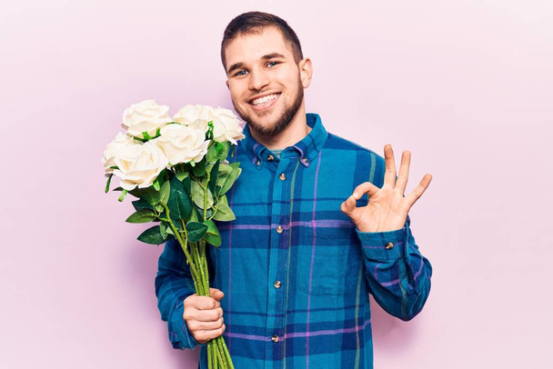 Νεαρός όμορφος άνδρας κρατώντας μπουκέτο λουλούδια κάνει ok υπογράψει με τα δάχτυλα, χαμογελώντας φιλικό gesturing εξαιρετικό σύμβολο  - Φωτογραφία, εικόνα