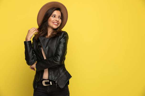 happy young woman in leather jacket looking to side and smiling, running fingers through hair on yellow background - Φωτογραφία, εικόνα