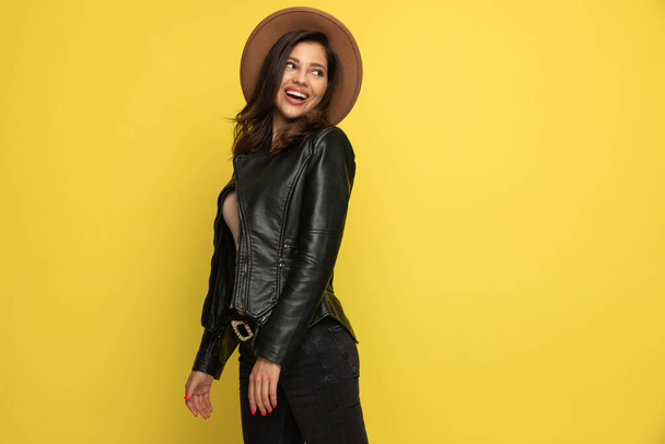 happy young woman in leather jacket looking to side and smiling on yellow background - Photo, Image