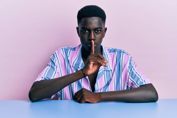 若いアフリカ系アメリカ人の男性は、テーブルの上に座っているカジュアルな服を着て、唇に指で静かになるように求めています。沈黙と秘密の概念.  - 写真・画像